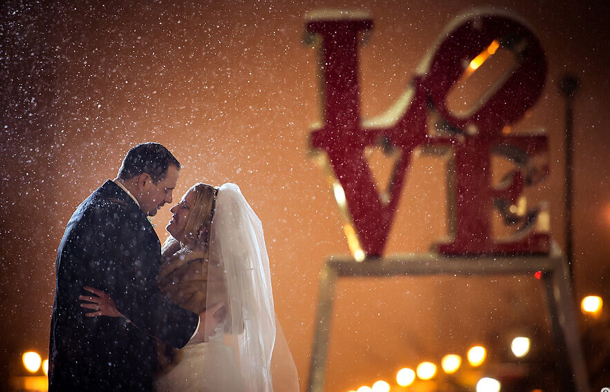 A bride and groom standing in front of a love sign in snowy New York City, capturing their beautiful winter wedding in the heart of NYC.