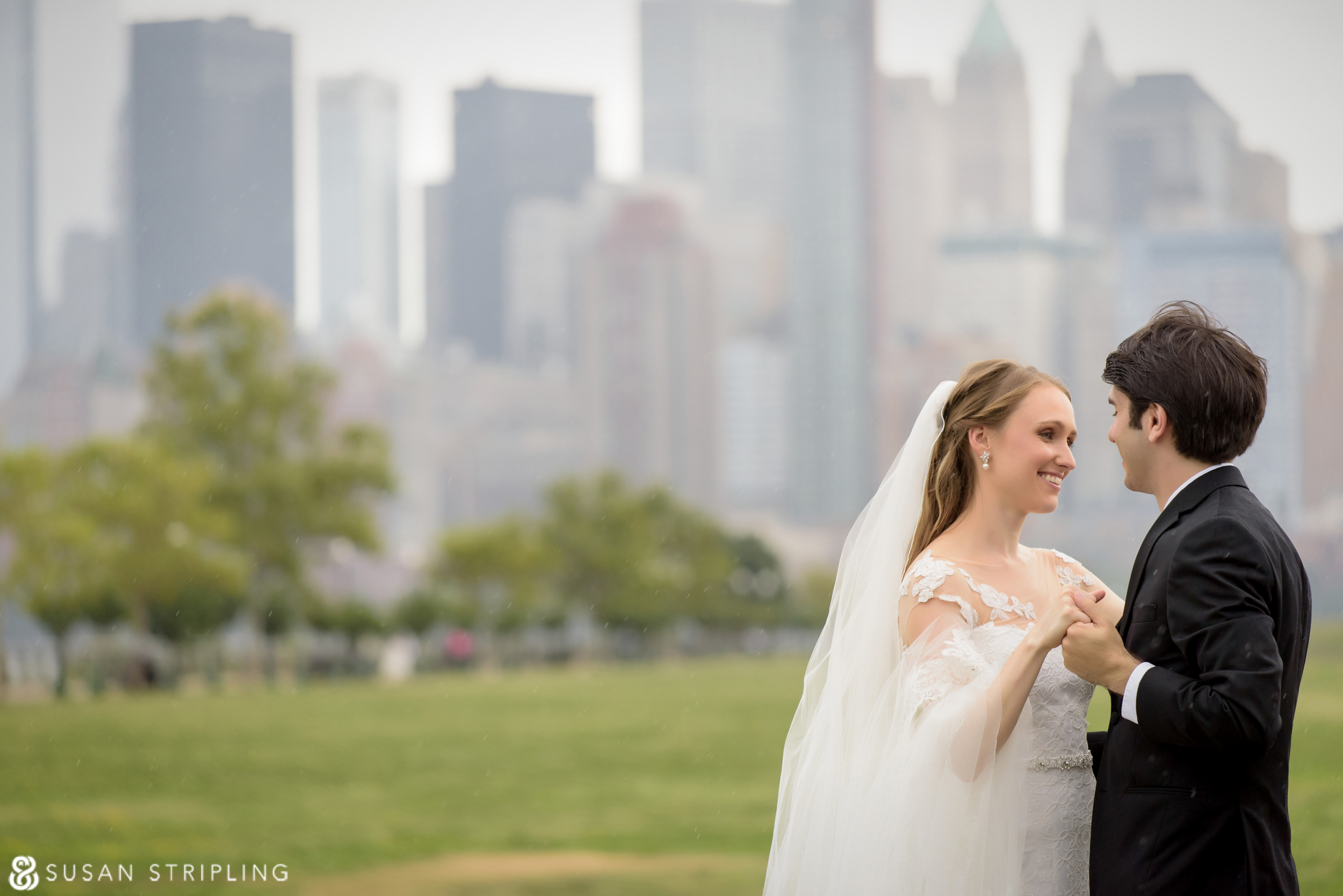 A bride and groom hold hands and smile at each other outdoors, with a city skyline and greenery in the background—a perfect highlight from their 2016 year in review.