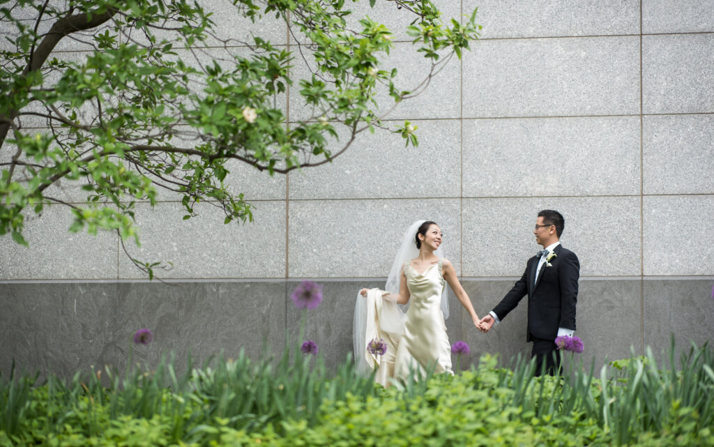 A bride and groom joyfully holding hands in front of the elegant Ritz Carlton Battery Park, commemorating their wedding day.