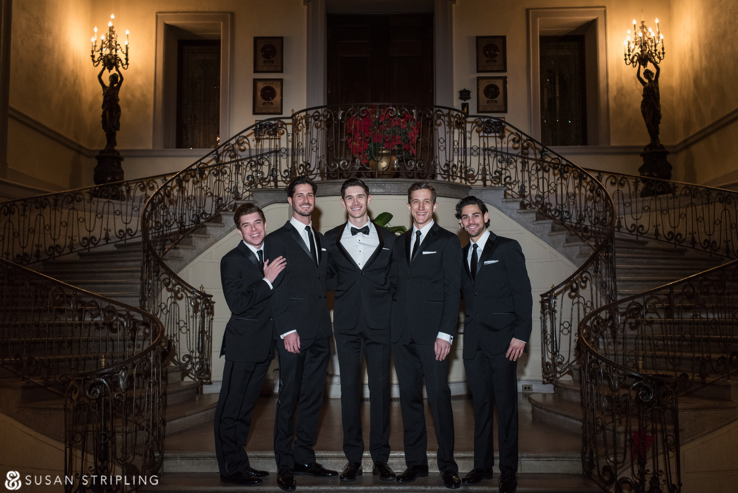 A group of groomsmen in tuxedos posing in front of a staircase during family pictures at a wedding.