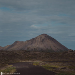 A woman is walking down a dirt path with a mountain in the background, resembling a Game of Thrones location.