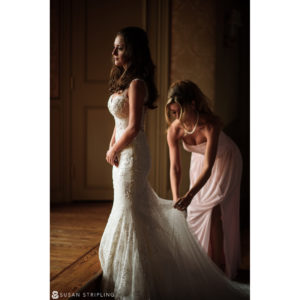 Two women assisting each other during an outdoor wedding at Oheka Castle as they put on a wedding dress together.