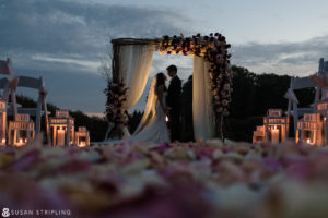 A bride and groom share a romantic kiss under a candlelit arch at their outdoor wedding at Oheka Castle.