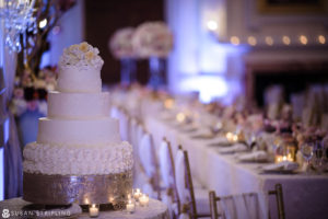 A beautiful wedding cake sits on a table in an elegant room at the Oheka Castle, surrounded by the lush greenery of an outdoor wedding ceremony.