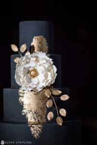 A black and gold wedding cake with a flower on top, perfect for a Cescaphe Philadelphia wedding.