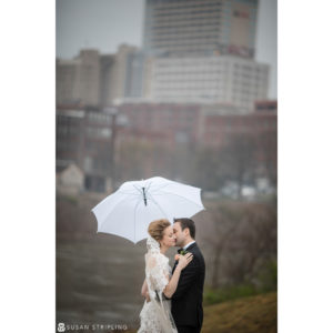 A destination wedding photographer captures a picturesque moment as a bride and groom share a romantic kiss under an umbrella amidst the backdrop of a bustling city.