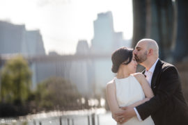 A summer elopement at Brooklyn Bridge Park with a bride and groom embracing in front of the iconic bridge.