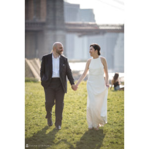 A summer elopement at Brooklyn Bridge Park, with a bride and groom holding hands in the grass.