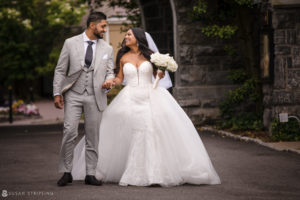 An Indian bride and groom holding hands in front of the historic stone building at Tarrytown House Estate during their wedding ceremony.