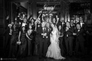 A black and white photo of a wedding party posing at the Crystal Tea Room, surrounded by confetti.