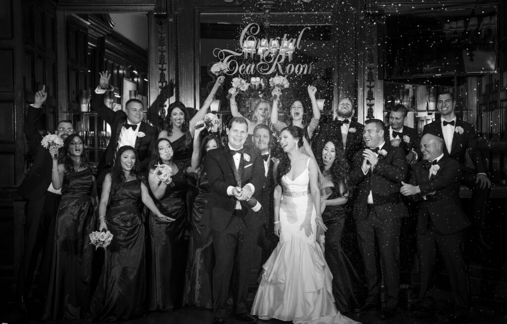 A black and white photo of a wedding party posing at the Crystal Tea Room, surrounded by confetti.