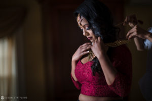 An Indian bride in a beautiful wedding dress putting on her necklace at Tarrytown House Estate.