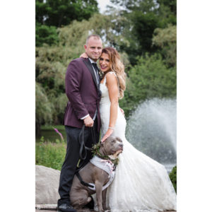 A bride and groom posing with their dog at a wedding celebration in front of a fountain.