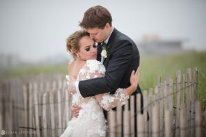 A bride and groom hugging at their wedding in front of a fence at the Bridgehampton Tennis and Surf Club.