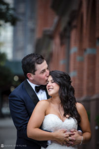 A bride and groom embracing in front of a brick building at their wedding.