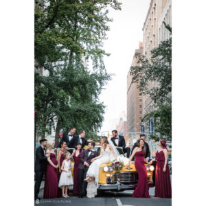 A group of bridesmaids and groomsmen pose in front of a yellow cab at a summer wedding.