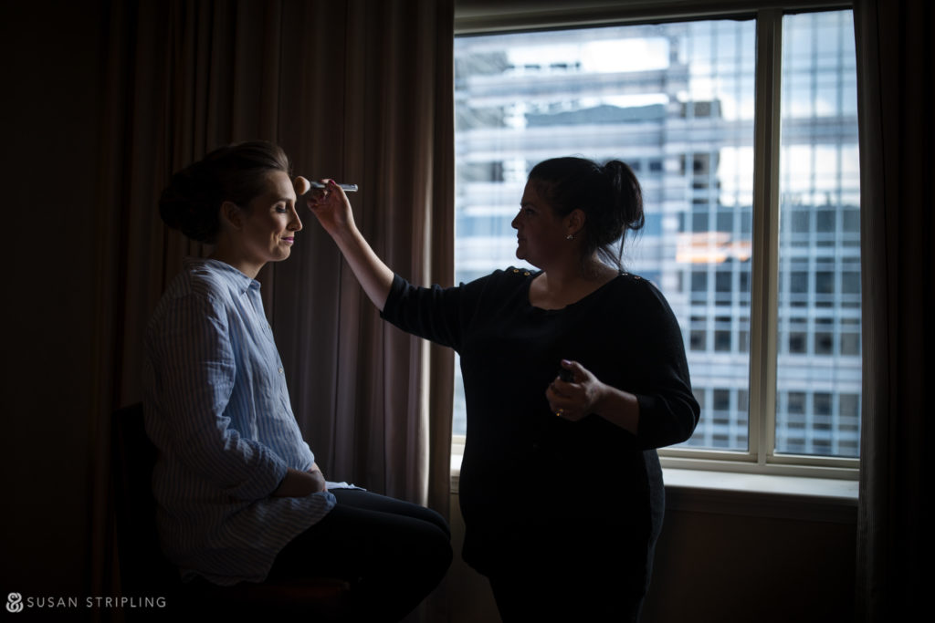 bride getting ready in dramatic windowlight with her makeup artist brushing powder on her cheeks