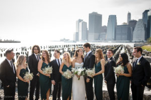 A group of bridesmaids and groomsmen pose for a wedding photo in front of the Manhattan skyline at 1 Hotel Brooklyn Bridge.