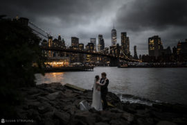 A bride and groom standing on rocks in front of the Brooklyn Bridge, capturing a beautiful elopement moment.