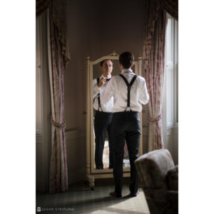 A man putting on suspenders in front of a mirror before attending a wedding at the Sleepy Hollow Country Club.