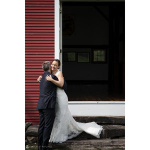 A bride and groom embracing in front of a rustic red barn at their charming riverside farm wedding.