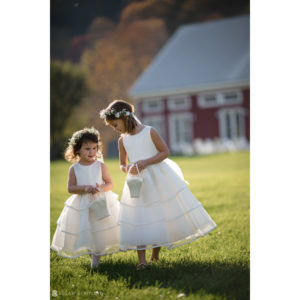 Two little girls in white dresses standing in front of a wedding barn.