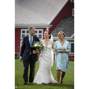 A bride and her mother walking in front of a rustic red barn at a picturesque riverside farm wedding.