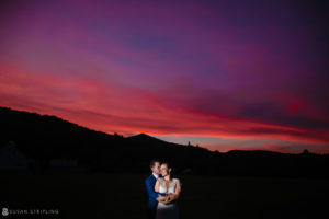 A bride and groom embrace in front of a colorful sunset during their riverside farm wedding.