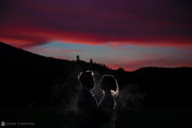 A silhouette of a bride and groom standing in front of mountains at sunset during their riverside farm wedding.