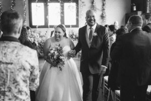 A bride walks down the aisle at her wedding, accompanied by her father.