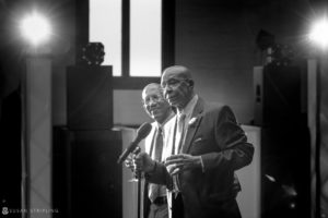 Two men in suits singing into microphones at a wedding.