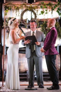 The beautiful wedding ceremony takes place at Quantum Leap Winery with the bride and groom exchanging vows in front of wine barrels.