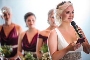 At the enchanting Quantum Leap Winery, a radiant bride stands surrounded by her bridesmaids, holding a microphone with excitement on her special wedding day.