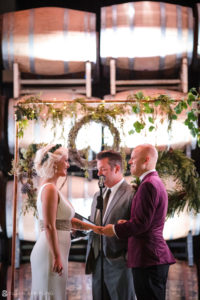 A bride and groom exchange vows at Quantum Leap Winery, surrounded by wine barrels.