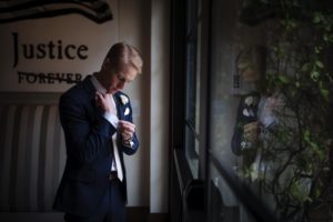 A man in a suit adjusts his tie in front of a window at a wedding venue, Alfond Inn.