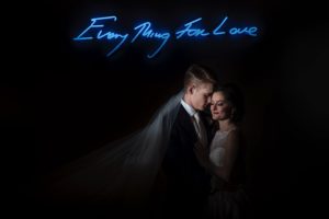 A wedding party, consisting of a bride and groom, posing in front of a neon sign that says "Every Man for Love" at the Alfond Inn.