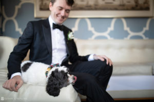 A man in a tuxedo sitting on a couch with a dog, surrounded by the elegance of Front and Palmer at a Winter Wedding.