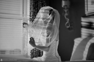 A bride, under the guidance of a Grand Master, elegantly adorns her veil while standing in front of a captivating window.