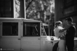 A bride and groom standing next to a vintage car outside Loews Hotel Philly on their wedding day.