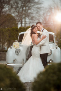 A bride and groom posing in front of a classic car at their wedding.
