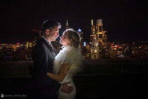 A wedding couple embracing in front of a city skyline at night at Loews Hotel Philly.