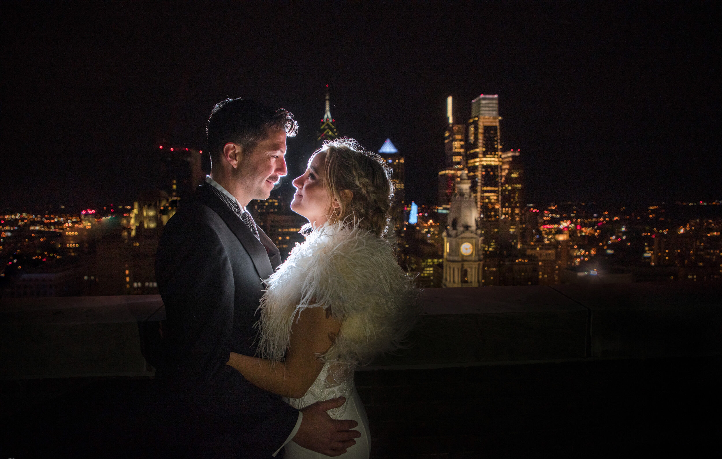A wedding couple embracing in front of a city skyline at night at Loews Hotel Philly.