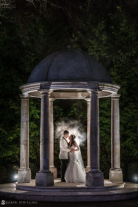 A bride and groom share a romantic kiss in front of a gazebo at the Florentine Gardens estate during their wedding at night.