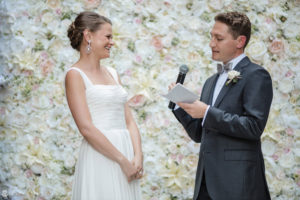 A summer bride and groom standing in front of a flower wall.