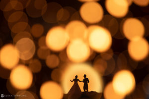 A romantic silhouette of a bride and groom standing in front of bokeh lights, capturing the enchanting essence of a summer wedding.