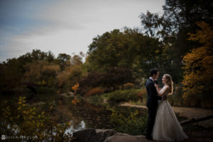 A bride and groom standing next to a pond in autumn at their wedding.