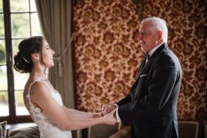 A bride and her father are holding hands in a room at Bourne Mansion during their summer wedding.