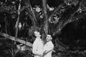 A black and white photo of two grooms on their wedding day, standing in front of a tree at the Ritz Carlton Dorado Beach.