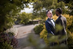 Two men in tuxedos standing in a summer wedding garden at Liberty Warehouse.