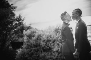 A black and white photo of two men kissing in front of bushes at a summer wedding at Liberty Warehouse.
